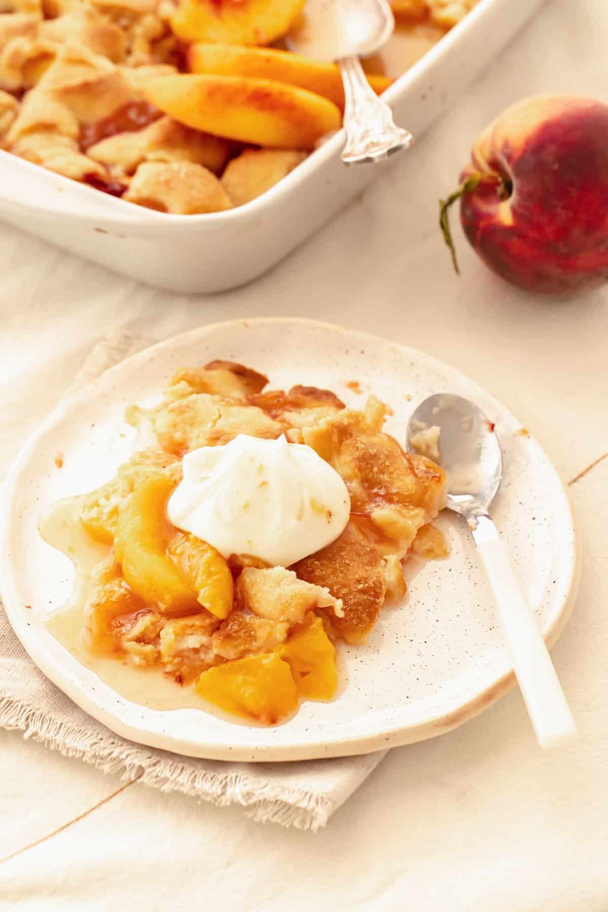 Serving of peach cobbler with whipped cream on white plate and surface. Partial view of baking dish in background. 
