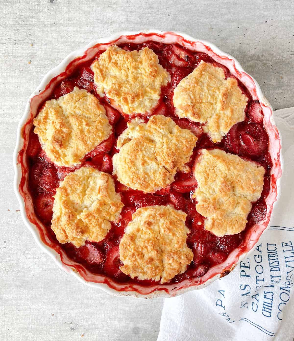 Round white dish with baked strawberry cobbler on grey surface, white kitchen towel.