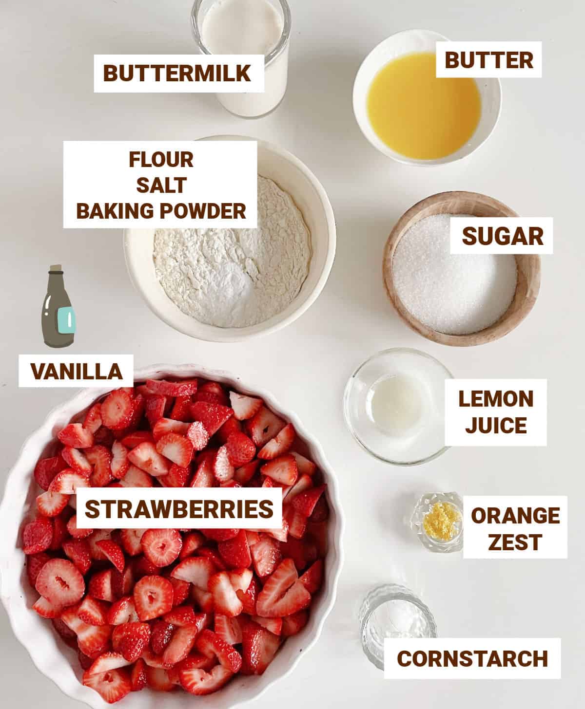 Grey surface with strawberry cobbler ingredients in bowls including lemon, butter, sugar, buttermilk, flour, cornstarch.