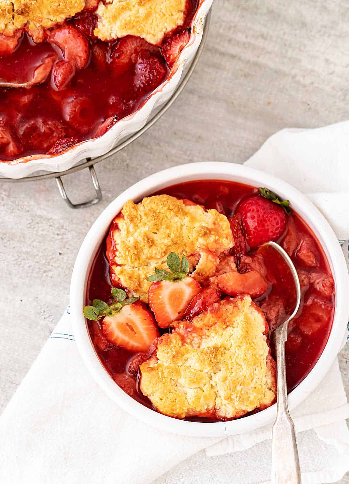 Serving of strawberry cobble in white bowl, white linen, silver spoon, dessert dish, on grey background.