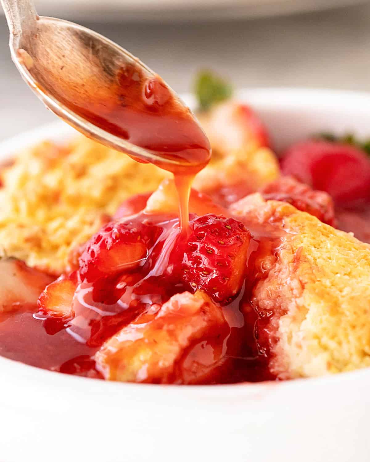 Silver spoon dripping stawberry syrup over serving of cobbler in white bowl.