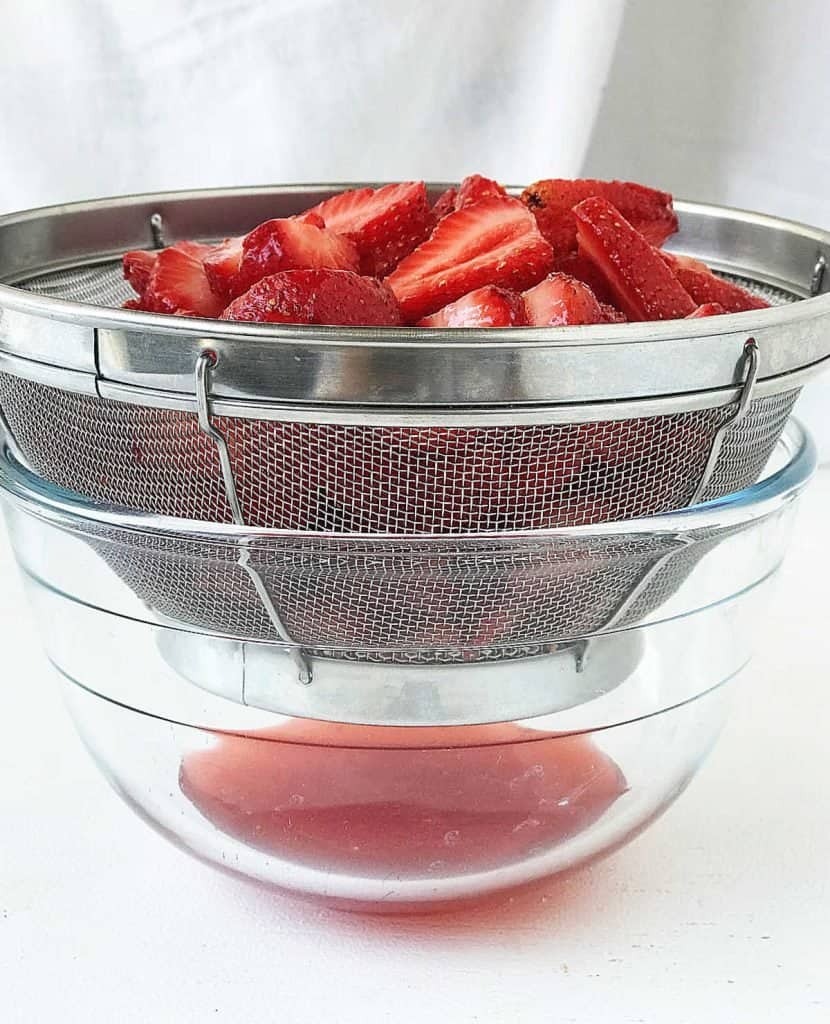 Metal colander draining strawberries over a glass bowl. White background.