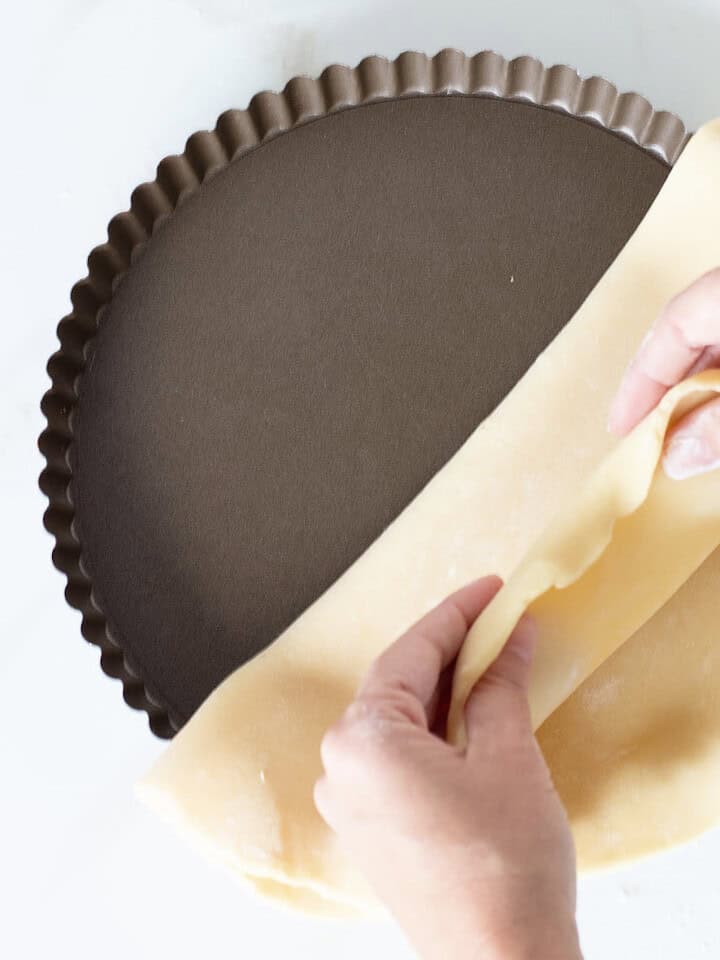 Hands lining a metal tart pan with pie crust. White surface. Top view.