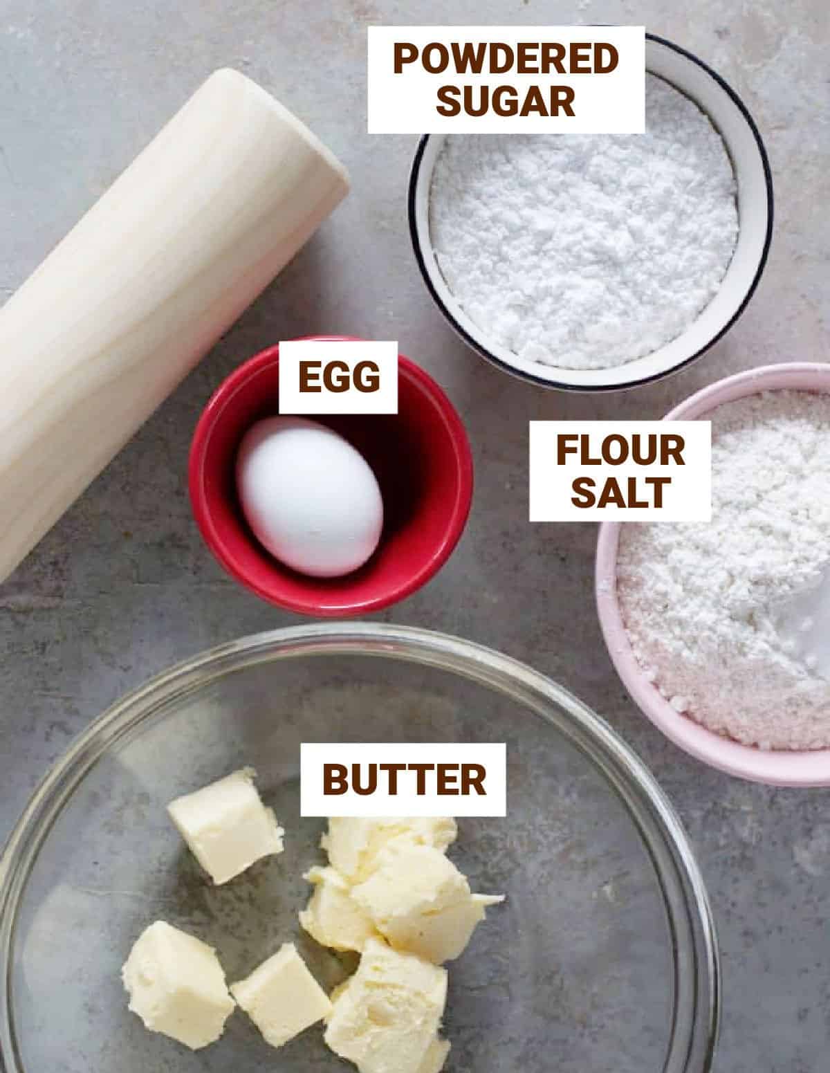 Sweet pie crust ingredients in bowls on grey surface, including butter, egg, sugar, salt, and rolling pin.