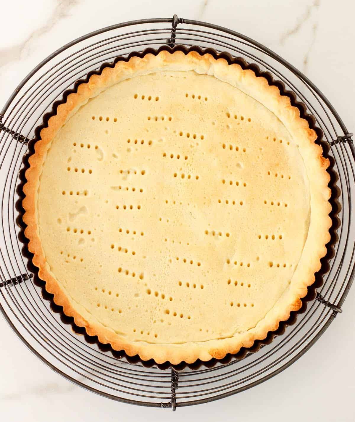 Baked shortcrust pastry on metal tart pan on wire rack, white marble surface.