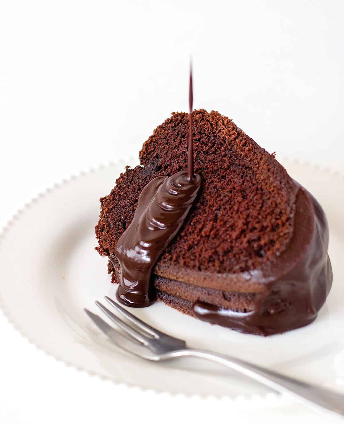 Slice of chocolate bundt cake on white plate, glaze pouring down.