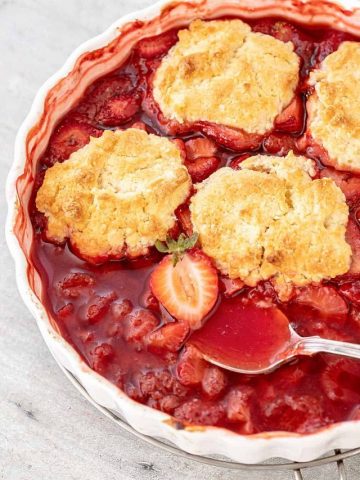 Close up of strawberry cobbler with silver spoon in white dish on grey surface.