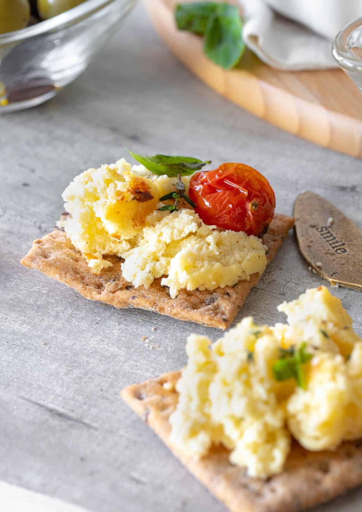 Crackers with baked ricotta, cherry tomato, and herbs on grey surface.