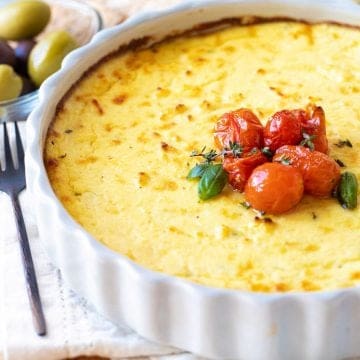 Grey round dish with baked ricotta topped with tomatoes, white linen underneath, olives in background.