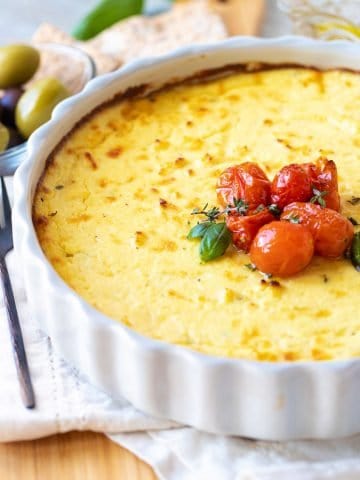 Grey round dish with baked ricotta topped with tomatoes, white linen underneath, olives in background.