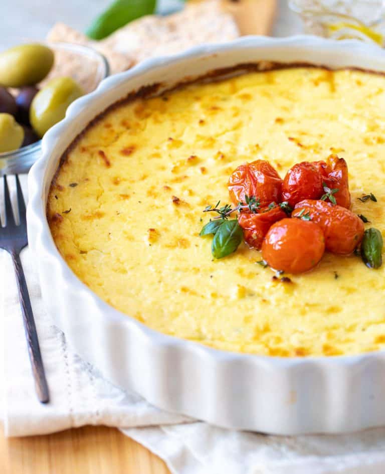 Grey round dish with baked ricotta topped with tomatoes, white linen underneath, olives in background.