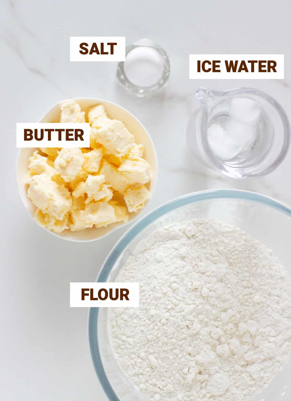 Basic shortcrust pastry ingredients in bowls on white surface including butter, flour, salt, water.