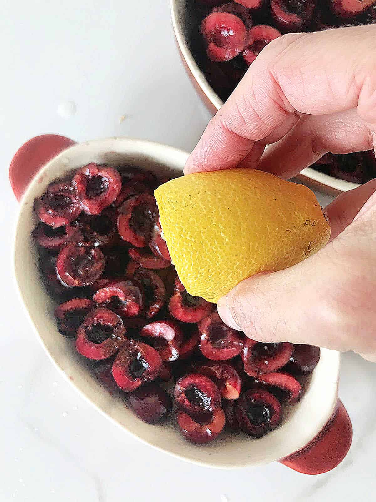 Hand adding lemon juice to fresh cherries in oval dish on white surface.