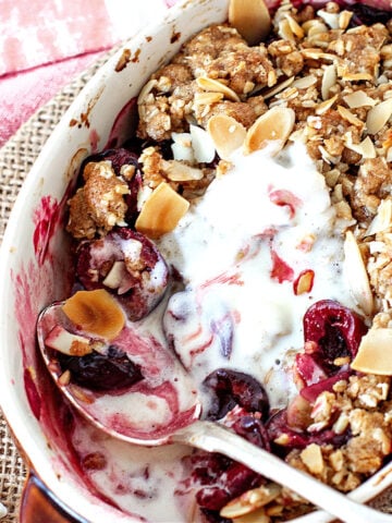 White individual dish with cherry crisp, silver spoon and melted ice cream, rustic background.