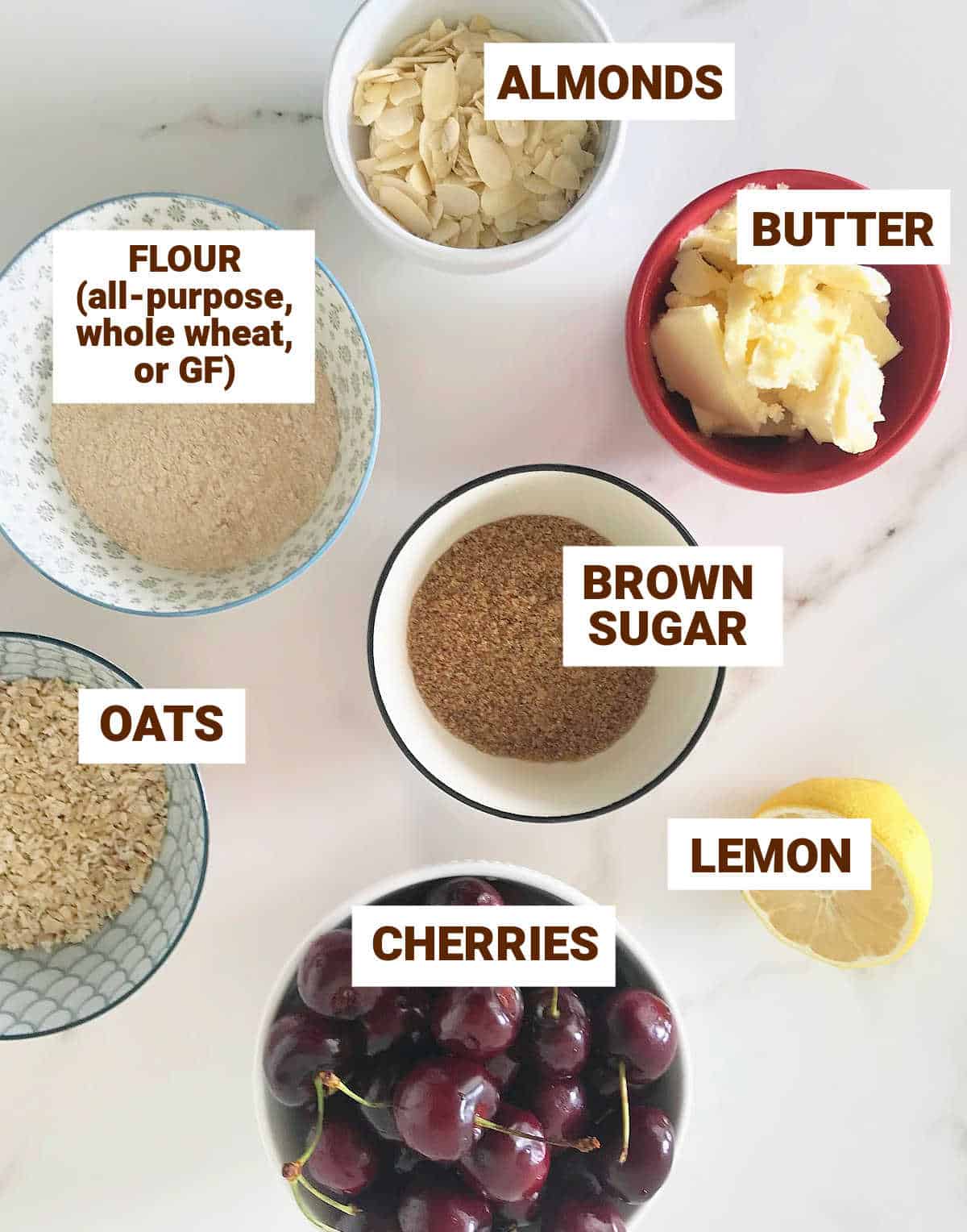 Colorful bowls on white surface with ingredients for almond cherry crisp including lemon, butter, brown sugar, oats.