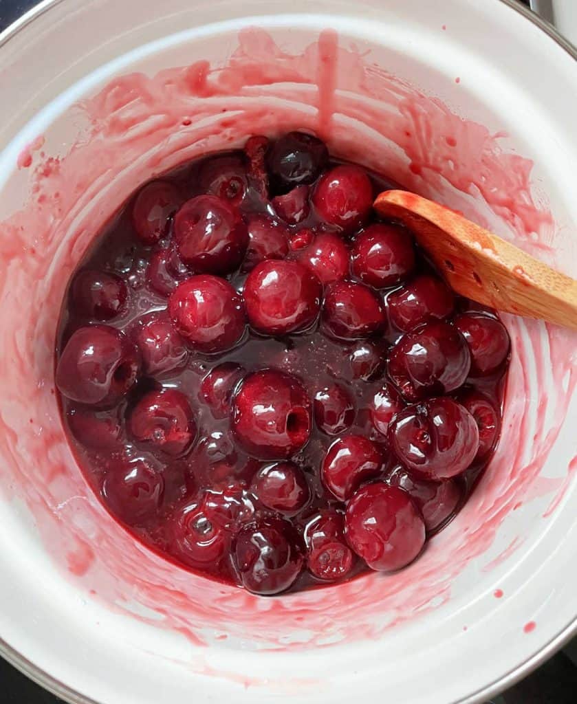 Top view of cherry pie filling simmering in white saucepan with wooden spoon.