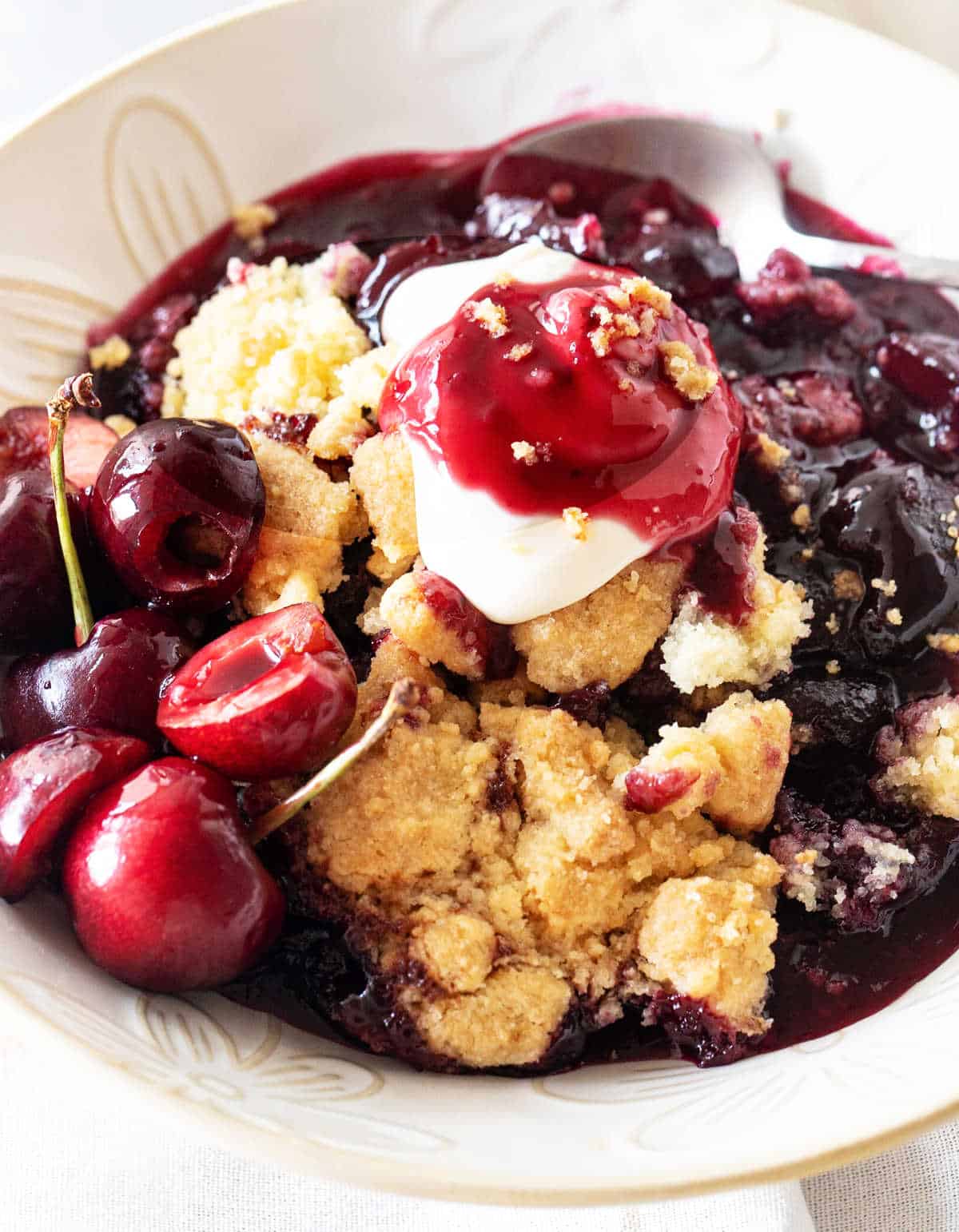 White bowl with cherry dump cake serving topped with whipped cream and fresh cherries.