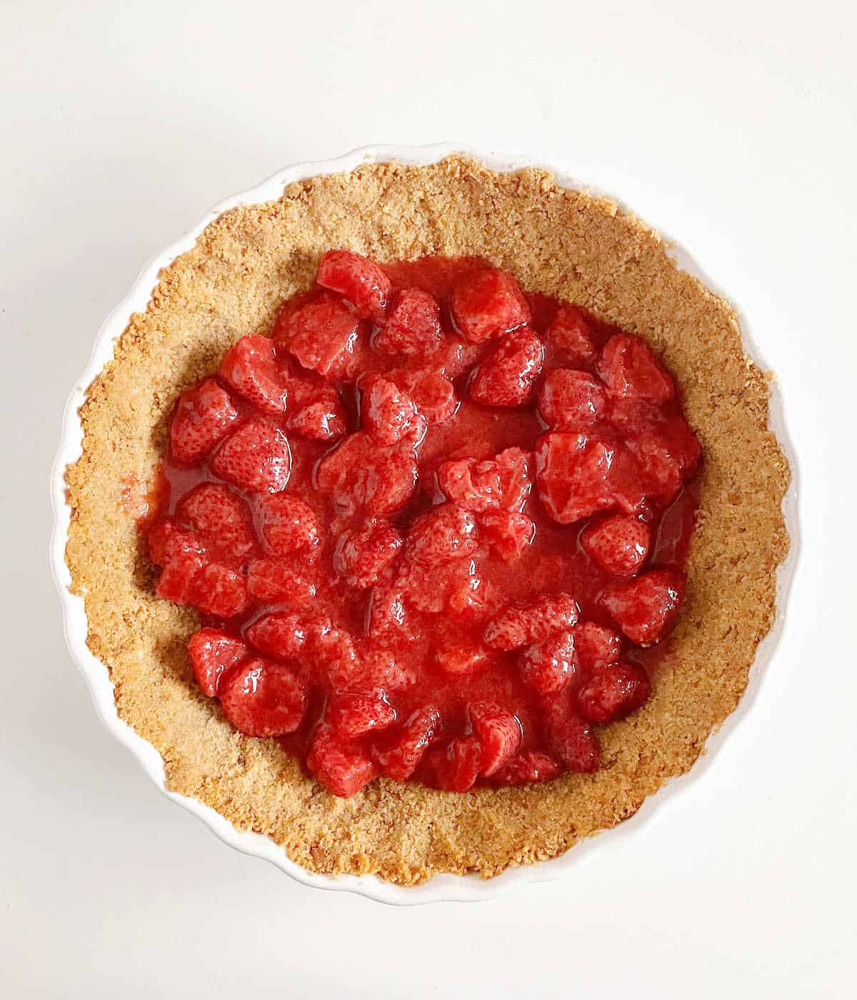 Top view of strawberry pie filling in a graham cracker crust on a white surface.