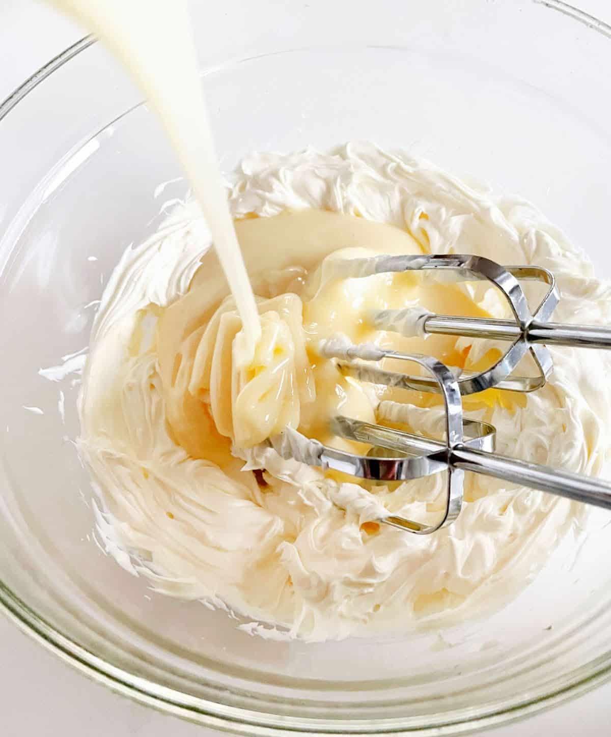 Pouring condensed milk cream cheese mixture in a glass bowl with electric beaters.