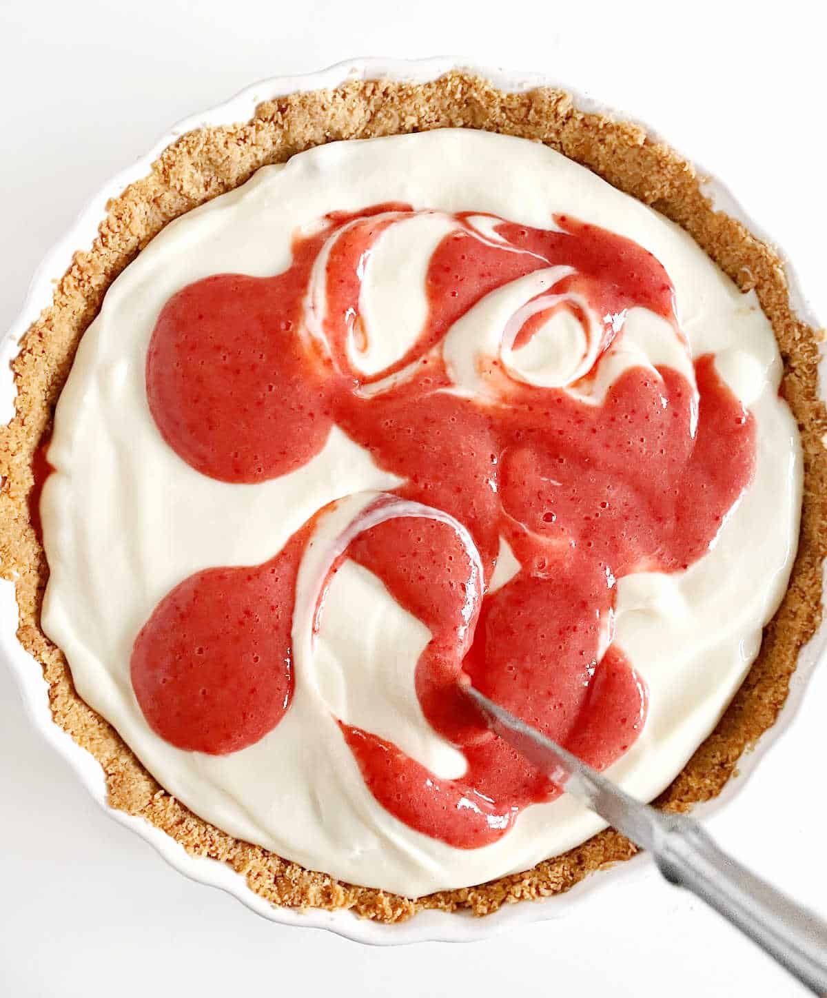 Swirling strawberry puree into cream cheese pie filling in a graham crust on a white surface.