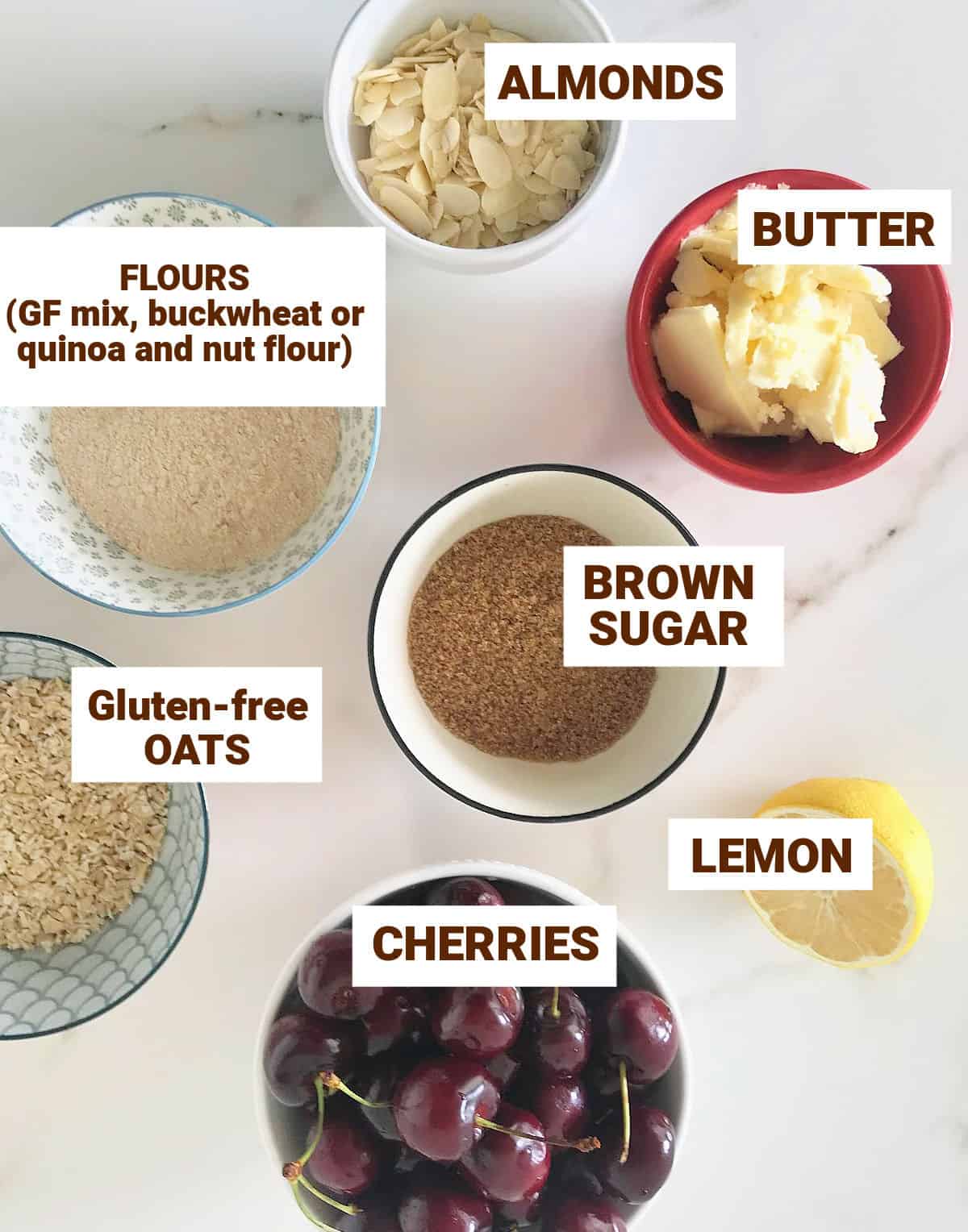 Colorful bowls on white surface with ingredients for almond cherry crisp including lemon, butter, brown sugar, oats.