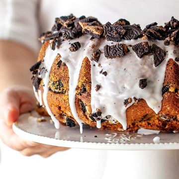 Hands holding glazed oreo bundt cake on a white cake stand.