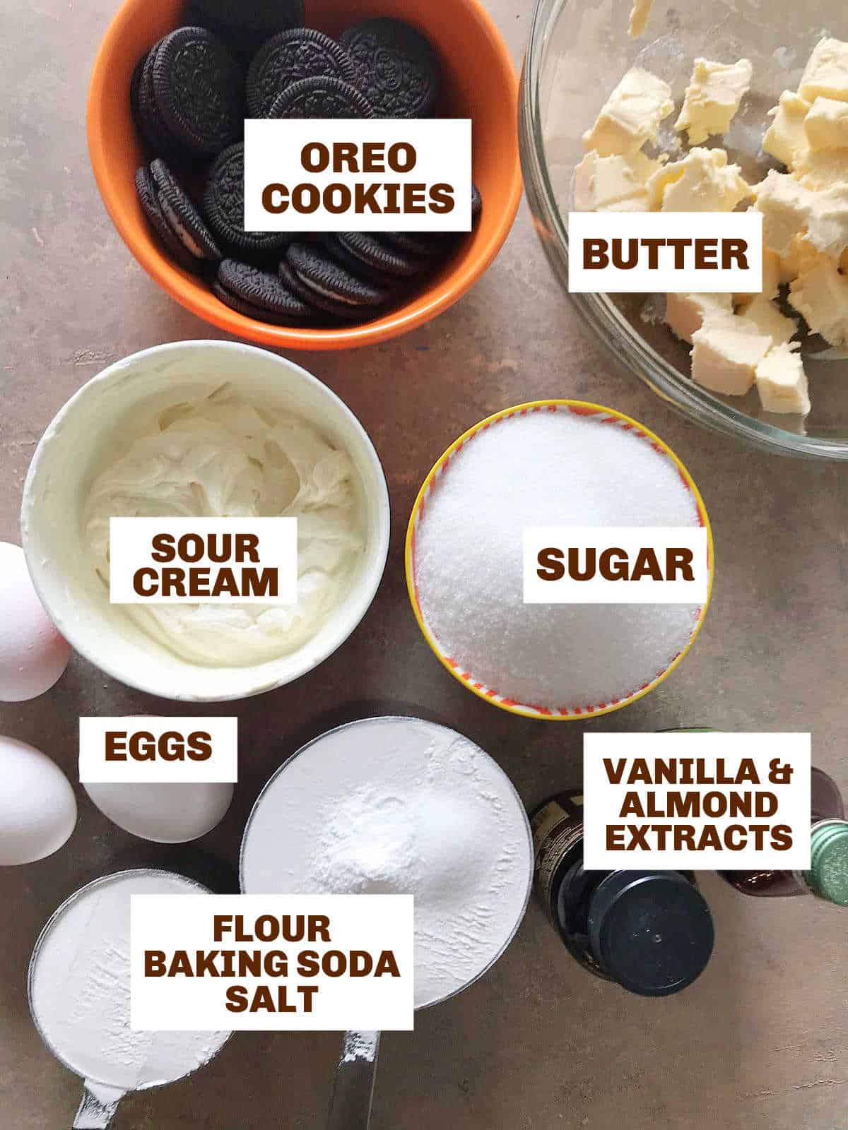 Brownish surface with bowls containing ingredients for oreo cake including butter, sugar, sour cream, flour, extracts, eggs.
