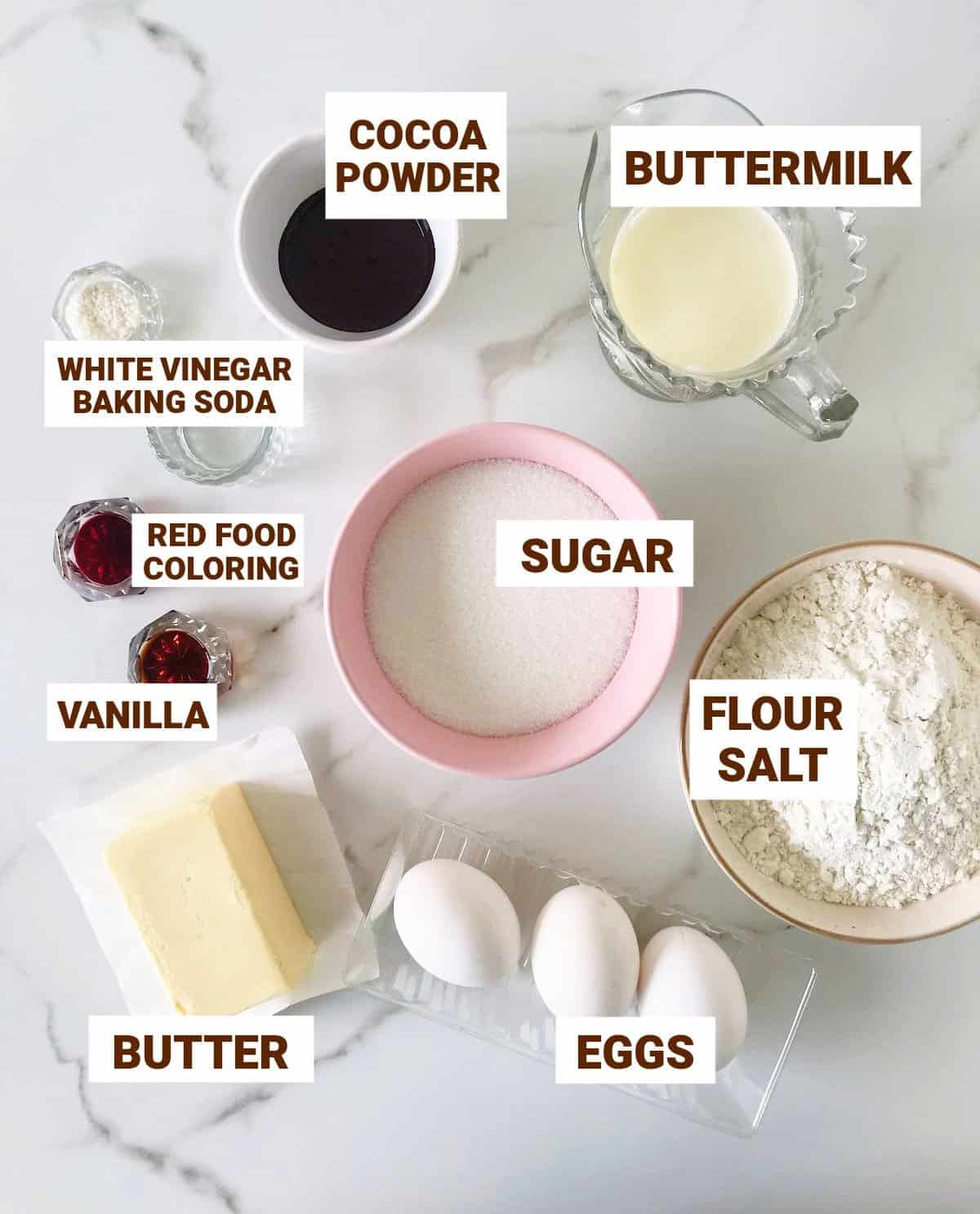 Red velvet cake ingredients in bowls on marble surface including butter, red coloring, buttermilk, vinegar, baking soda, cocoa powder.
