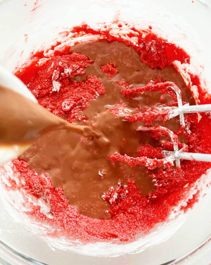 Adding chocolate milk to red cake batter in glass bowl. Metal beaters inside.