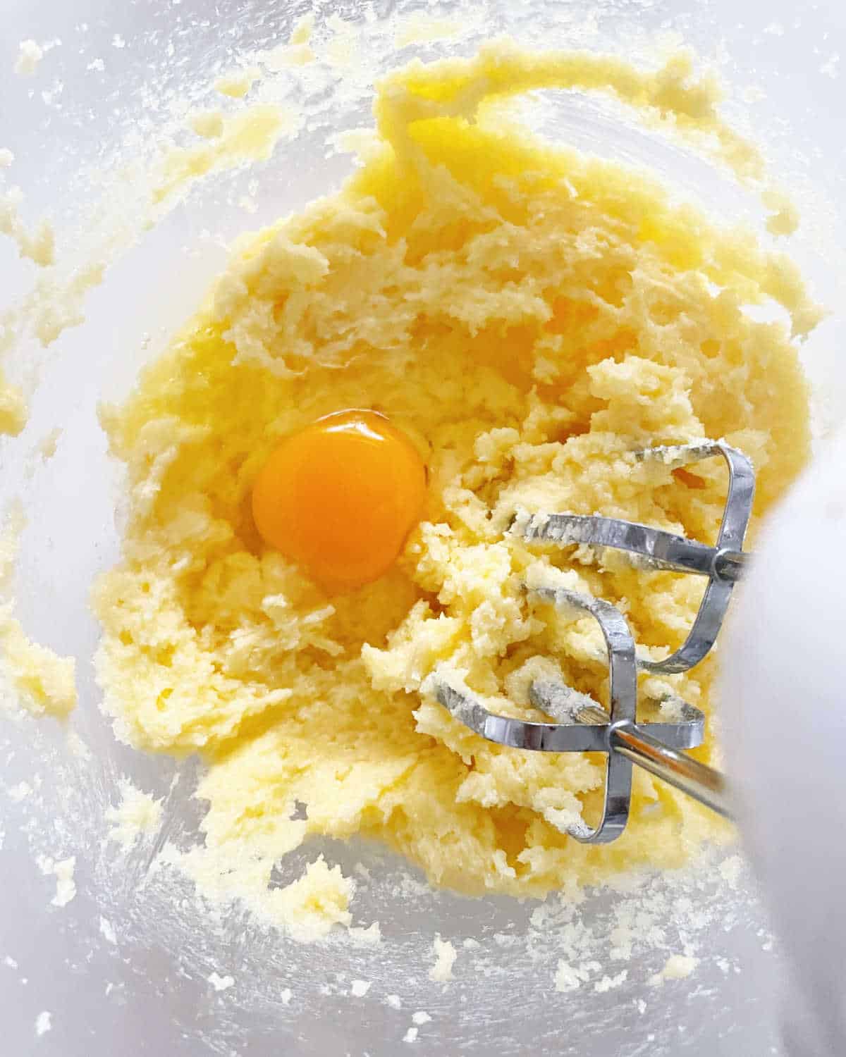 Butter and sugar mixed in glass bowl, one whole egg. White surface.