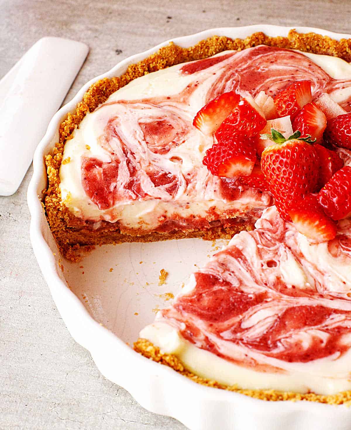 Strawberry pie missing a slice in white dish on grey surface, white cake server beside it. 