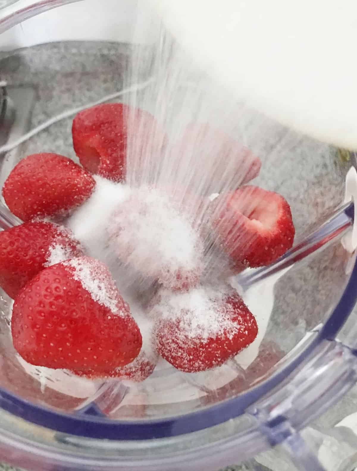 Adding sugar to whole strawberries in blender jar. 