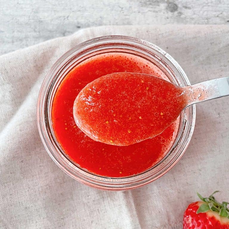 Glass jar and spoon with strawberry puree over a grey napkin.