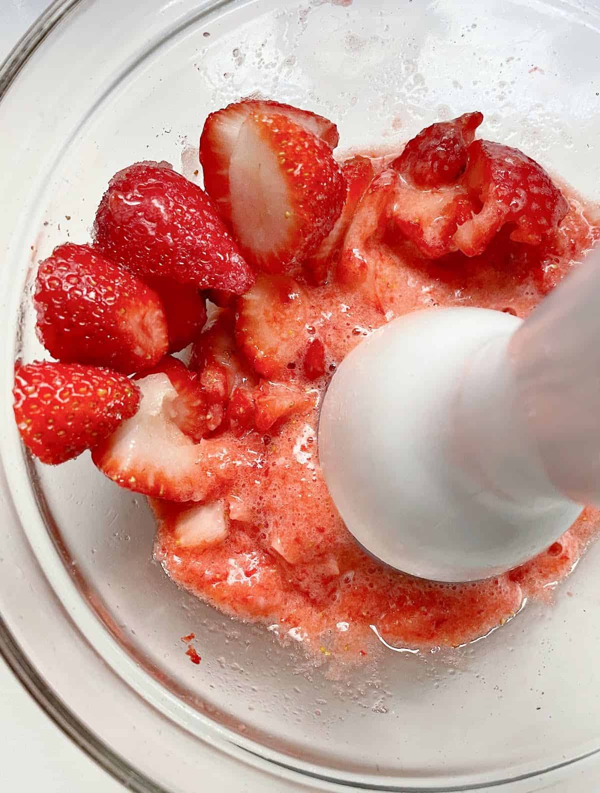 Immersion blender mixing semi whole strawberries with sugar in glass bowl.