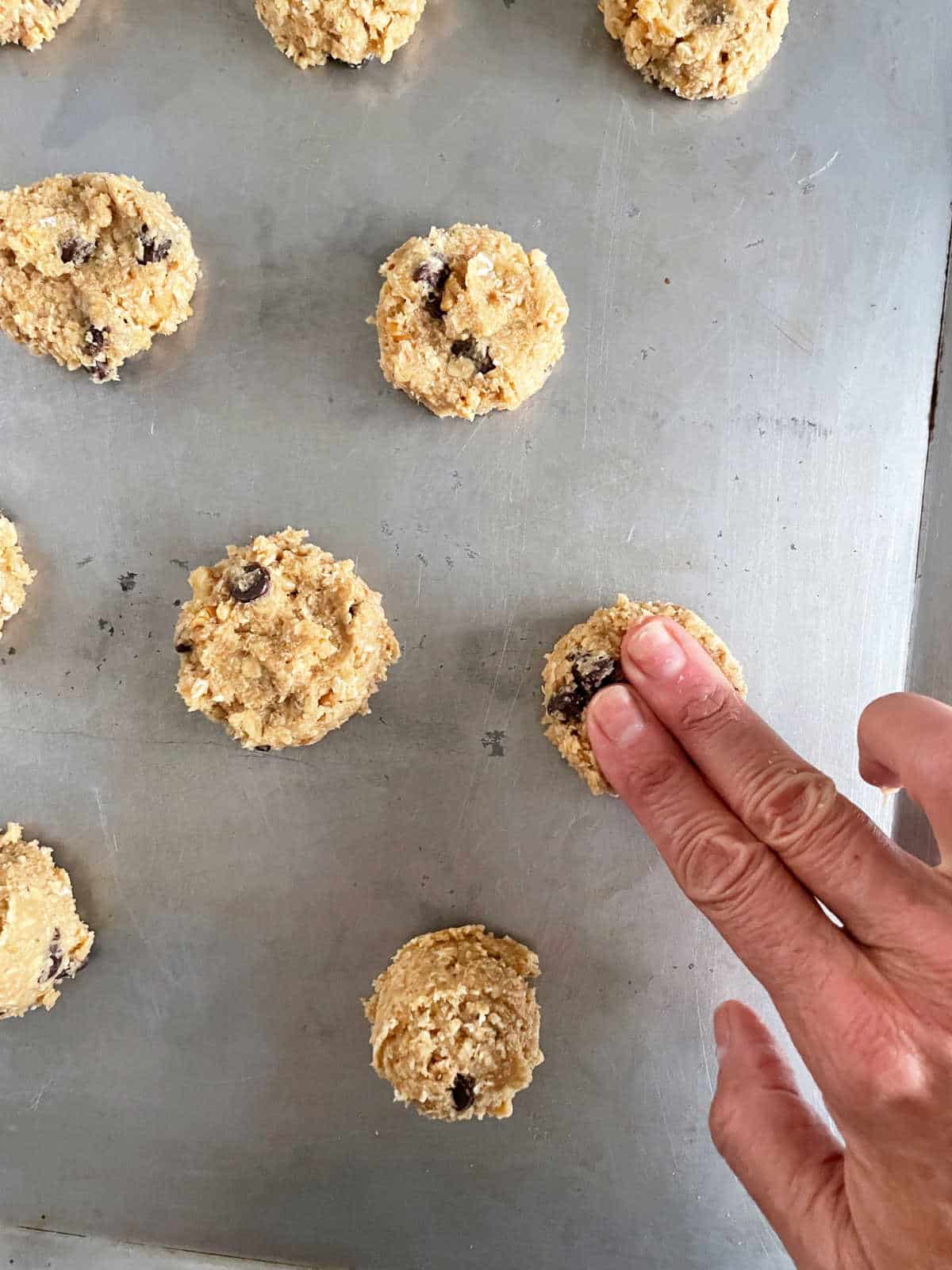 Hand pressing unbaked chocolate chip oatmeal cookies on metal cookie sheet.