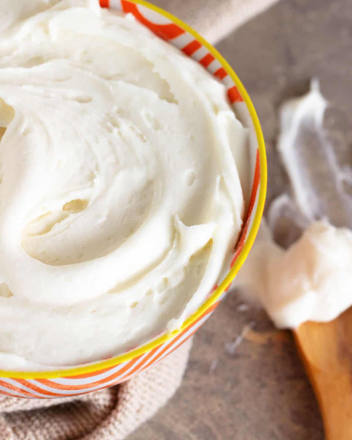 Striped orange bowl with cream cheese frosting, wooden spoon beside it, brownish surface.