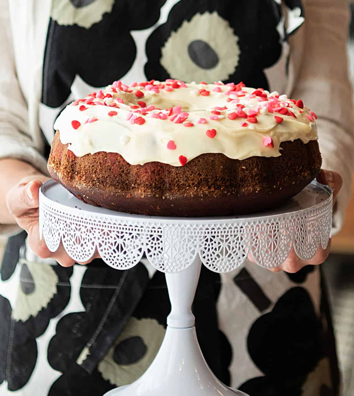 Partial view of person with apron holding frosted bundt cake on a white cake stand.