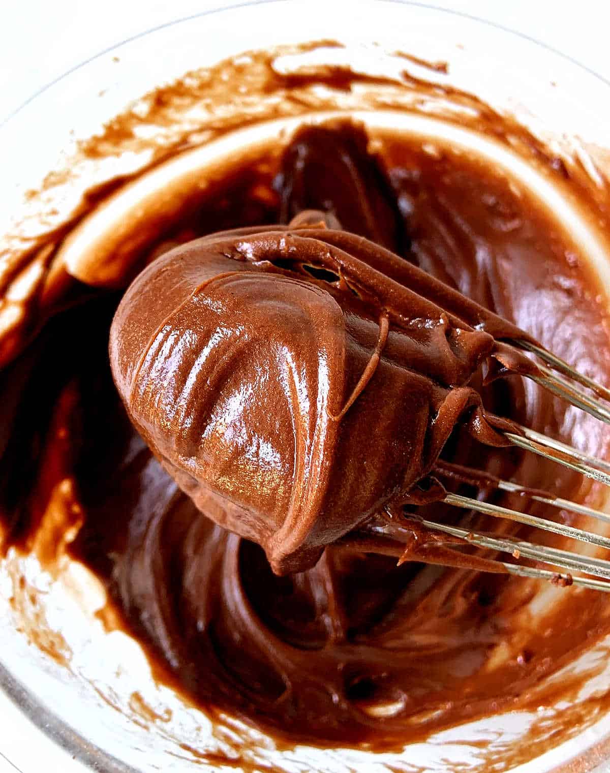 Shiny dollop of chocolate frosting on a wire whisk, bowl with more mixture below.