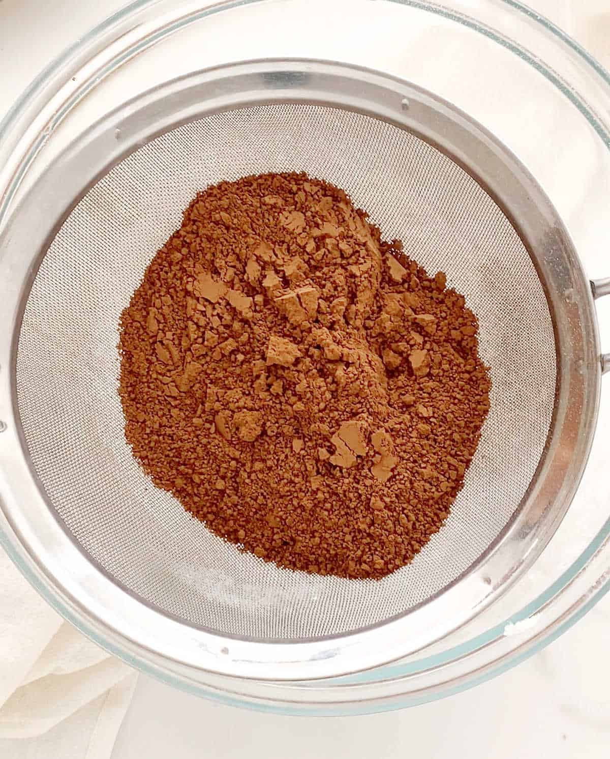 Sifting cocoa powder into a glass bowl on a white surface.