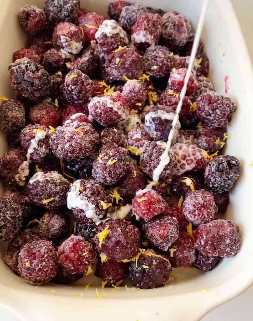 Pouring cornstarch mixture over blackberries with flavorings in a cream colored rectangular ceramic dish.