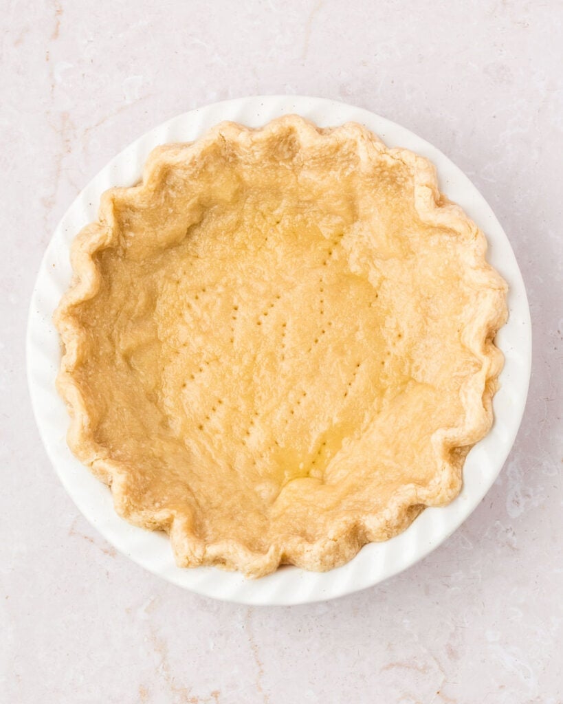 Pre-baked pie crust in a white dish. Top view. Light peach background.