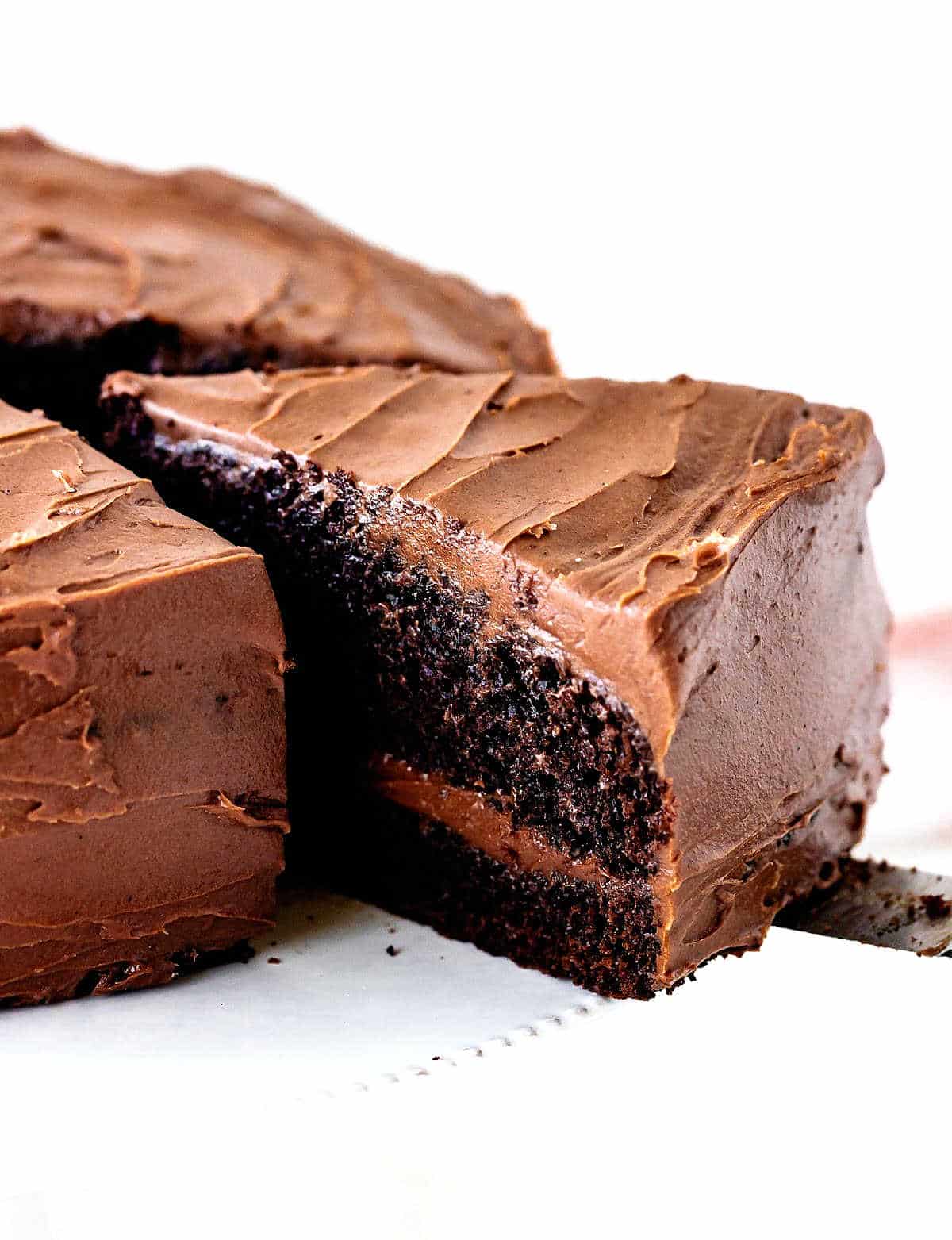 Removing a piece of filled and frosted chocolate cake on a white cake stand and background.