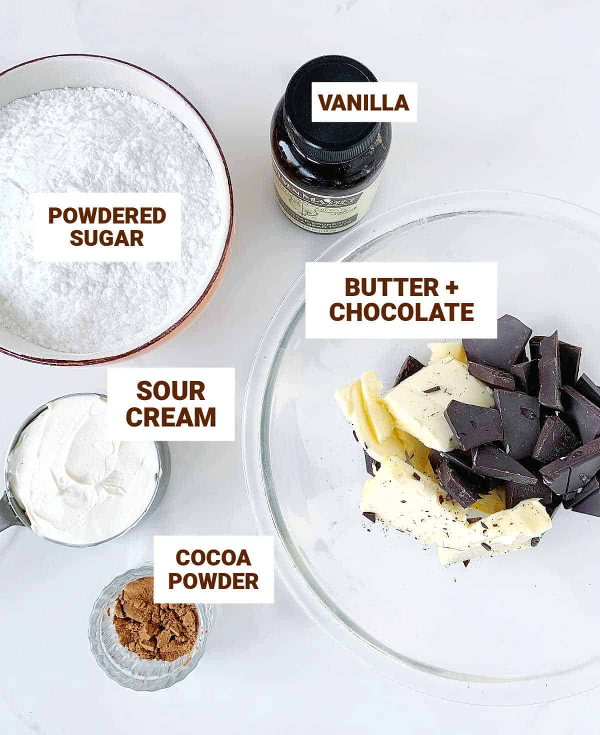 Bowls with ingredients for chocolate frosting including vanilla, cocoa powder, and measuring cup with sour cream.