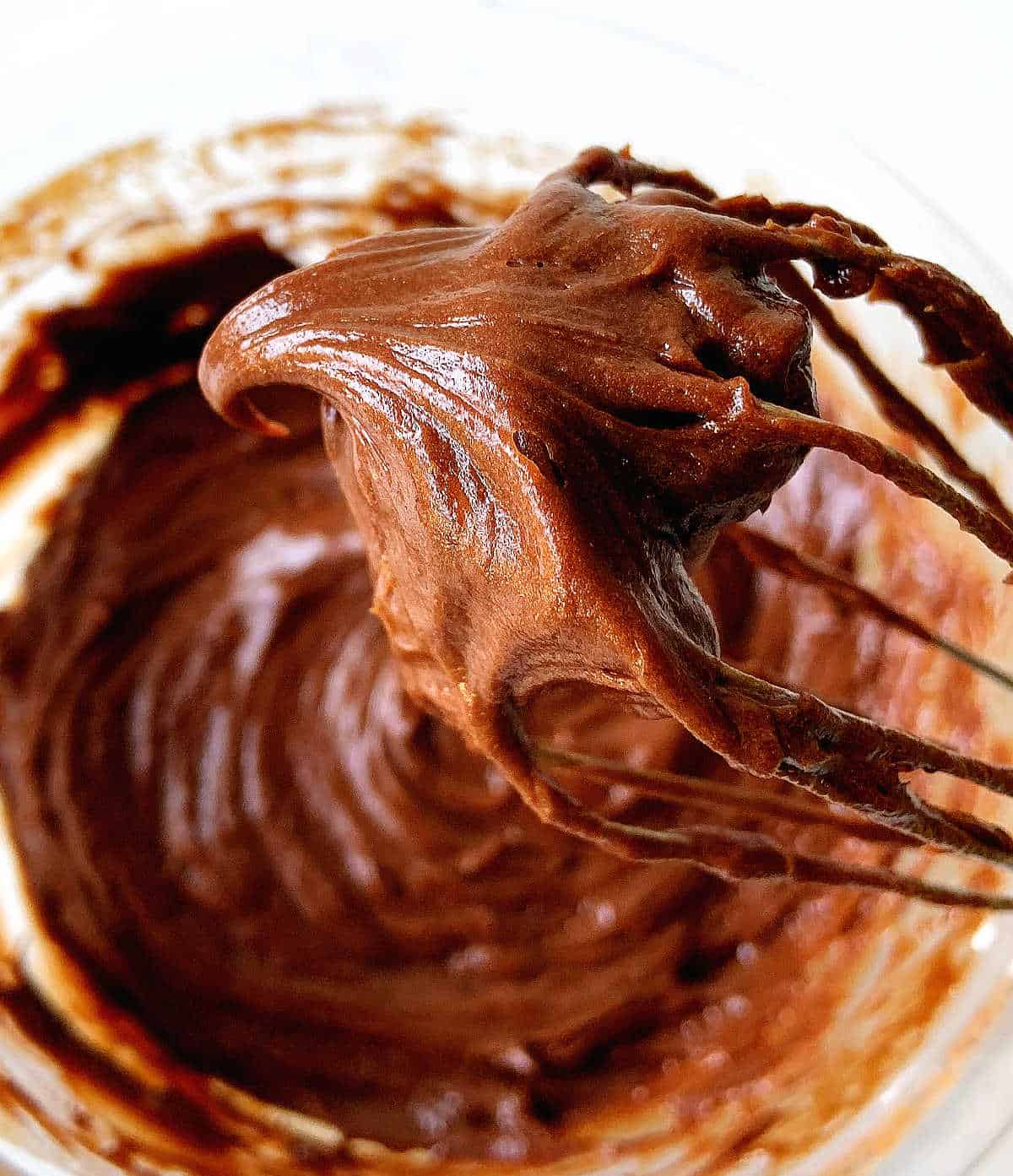 Wire whisk with dollop of chocolate frosting, glass bowl with rest of mixture below.