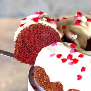 Close up of frosted red velvet bundt cake slice being pulled from whole cake. Beige and grey background.