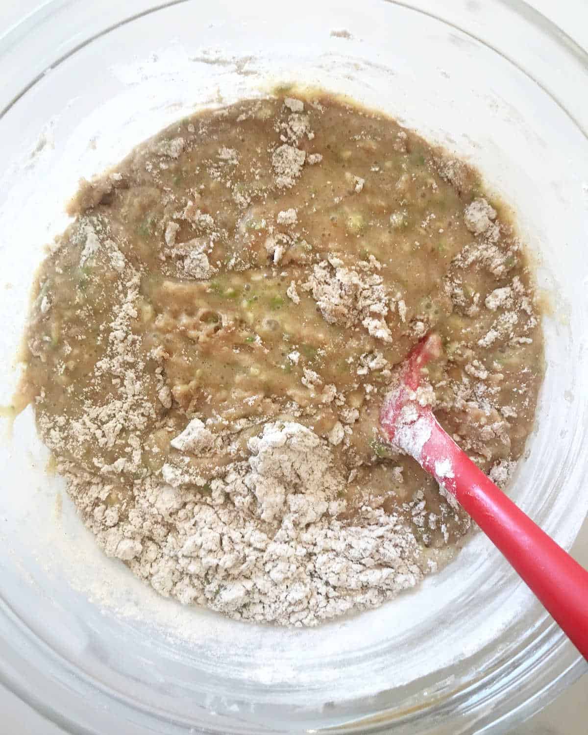 Mixing zucchini bread batter with whole wheat flour in glass bowl with red spatula.