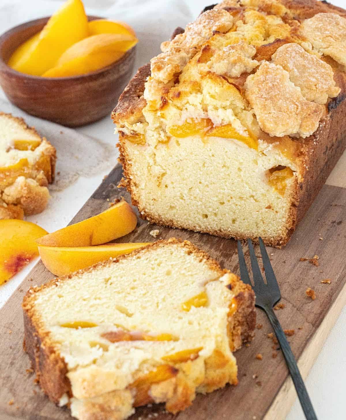 Peach pound cake with a cut slice on a wooden board, small dark fork and peach slices around.