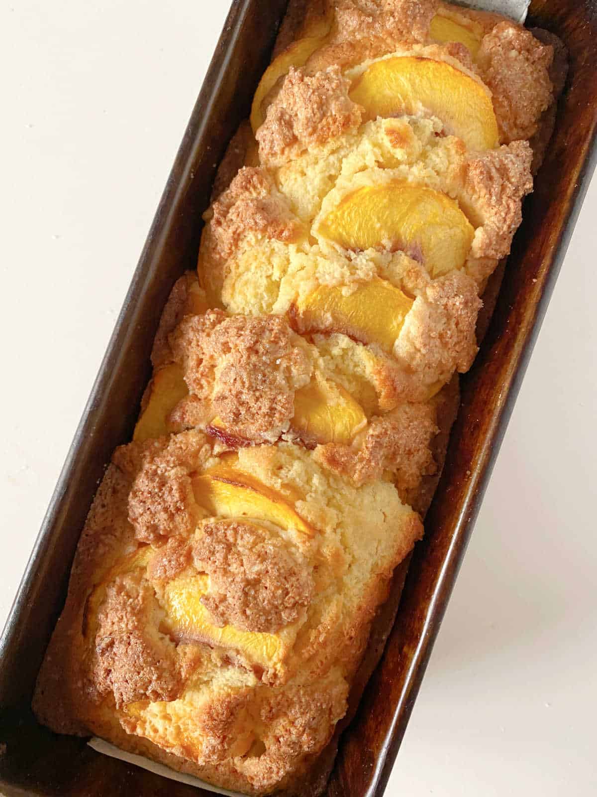 Partial top view of baked peach cake in dark metal loaf pan on a white surface.