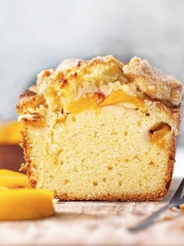 Front view of cut peach pound cake on wooden board. Grey background, peach slices, dark fork around.