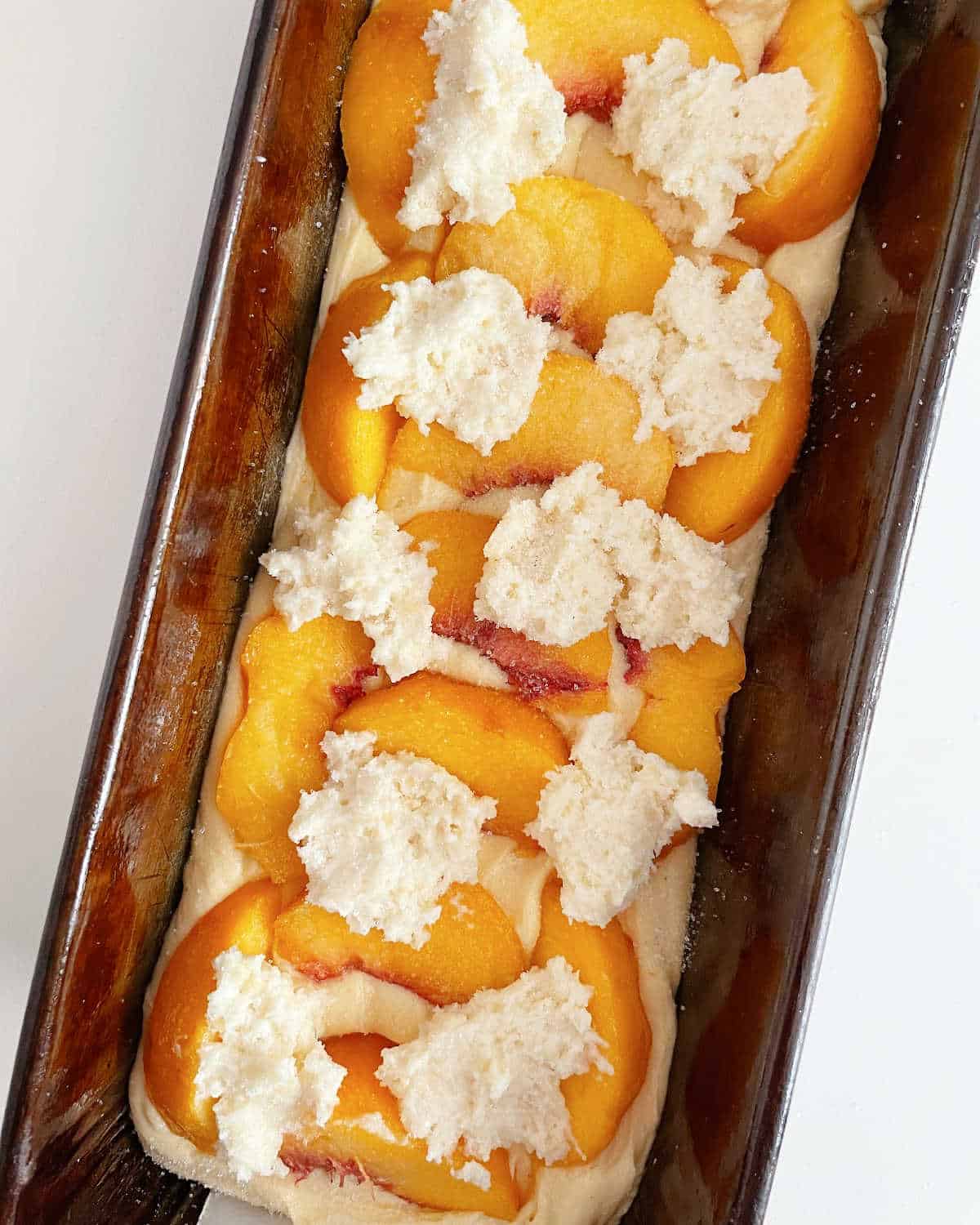 Unbaked peach pound cake with biscuit topping in metal pan on a white surface.